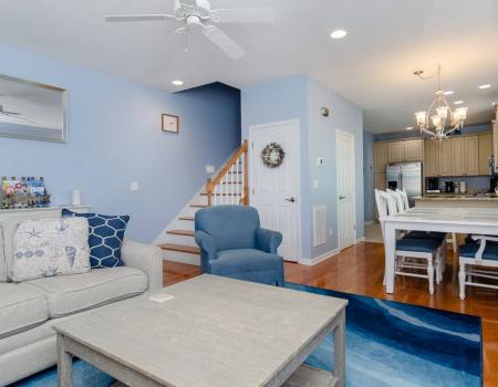 The cozy living room at Retreat Yo Self in Carolina Beach features blue carpet, blue walls, and a blue armchair with a white soft and coffee table. In the background, a staircase leads to an upper floor.