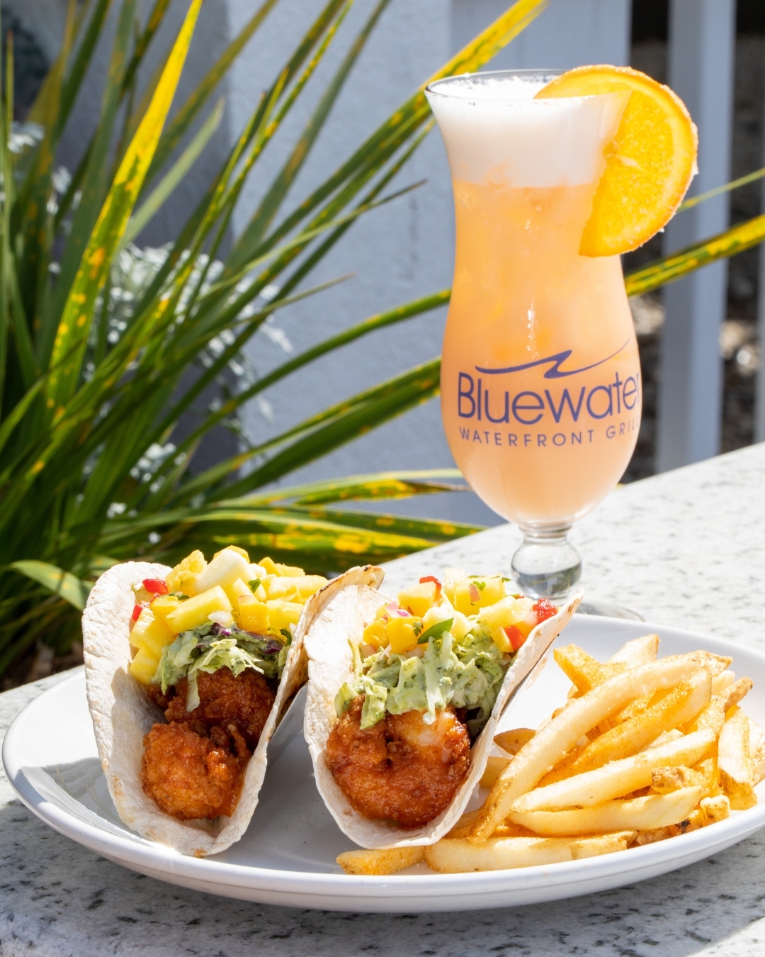 white plate with two fish tacos and fries in front of a citrus cocktail in a flute glass. credit: Bluewater Grill Instagram